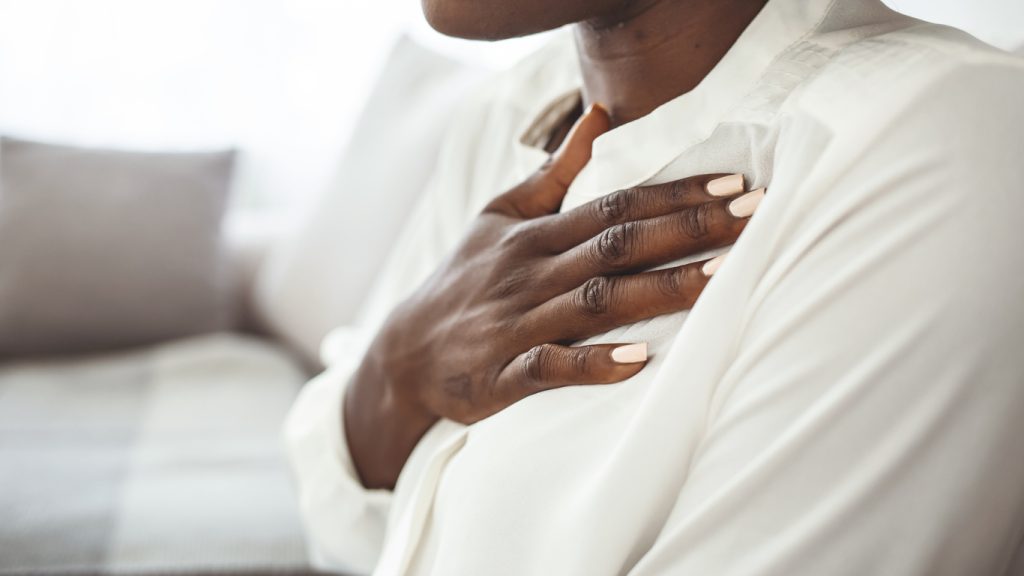 Girl having respiration problems touching chest sitting on a couch in the living room at home. Woman feeling pain ache touching chest having heart attack, sad worried lady suffers from heartache at home.