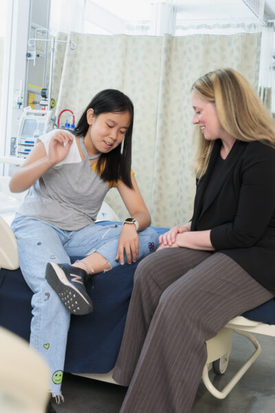 young girl sitting on hospital bed looking at watch on wrist. Beside her is a blonde woman also smiling and looking at watch on wrist.